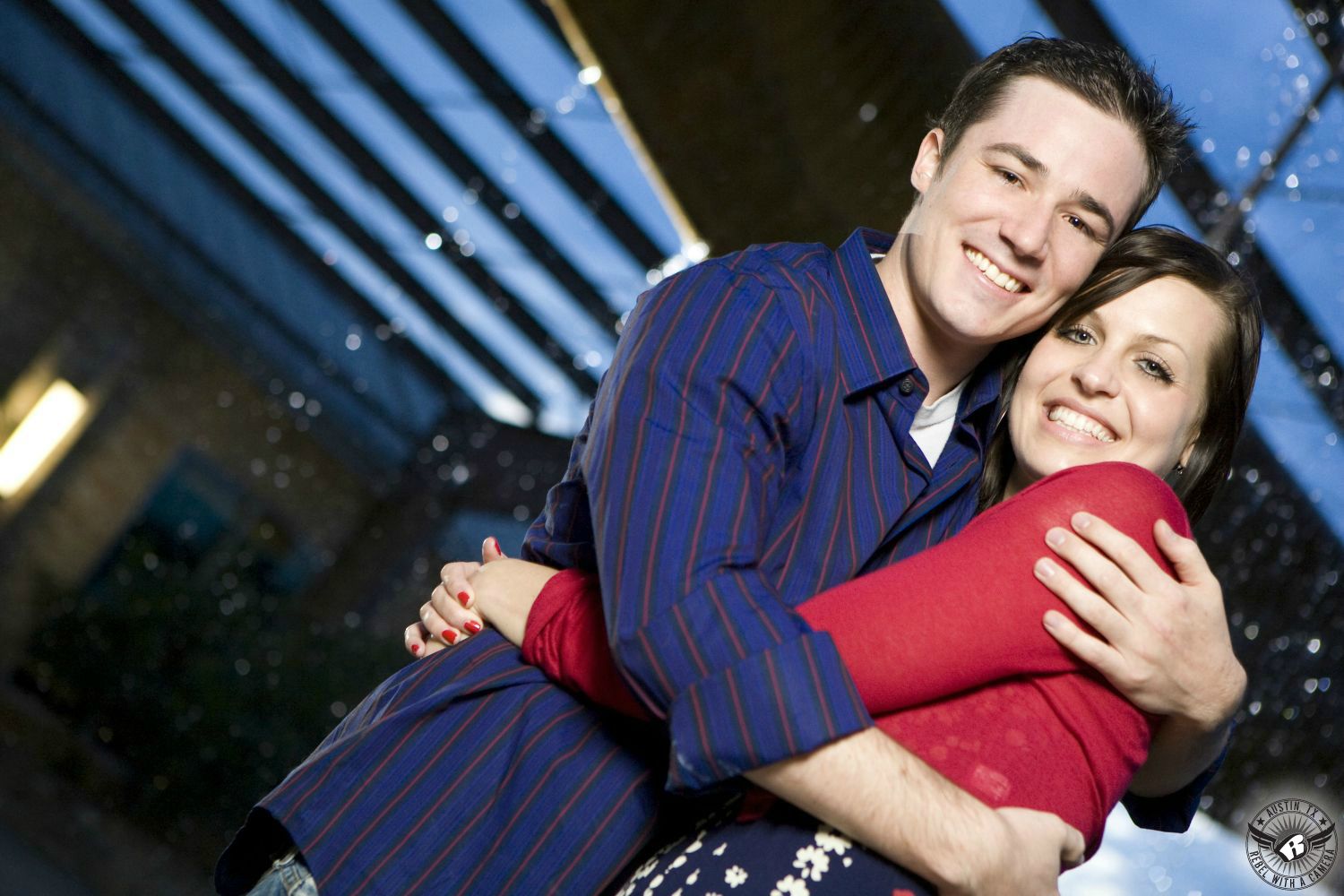 Delightful brunette lady wearing a sheer red blouse and a flower print dress is entwined with a dark spiky haired guy wearing a blue and orange button up collared shirt with the sleeves rolled up and blue jeans happily standing in front of a backlit fountain at night with light glinting off of drops of water in the background in this very Austin engagement picture in South Austin. 